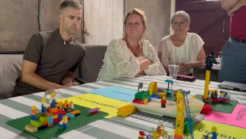 Drie deelnemers aan de partnerdag luisteren aan tafel naar de uitleg bij het gezamenlijke bouwwerk, dat voor hen op tafel staat