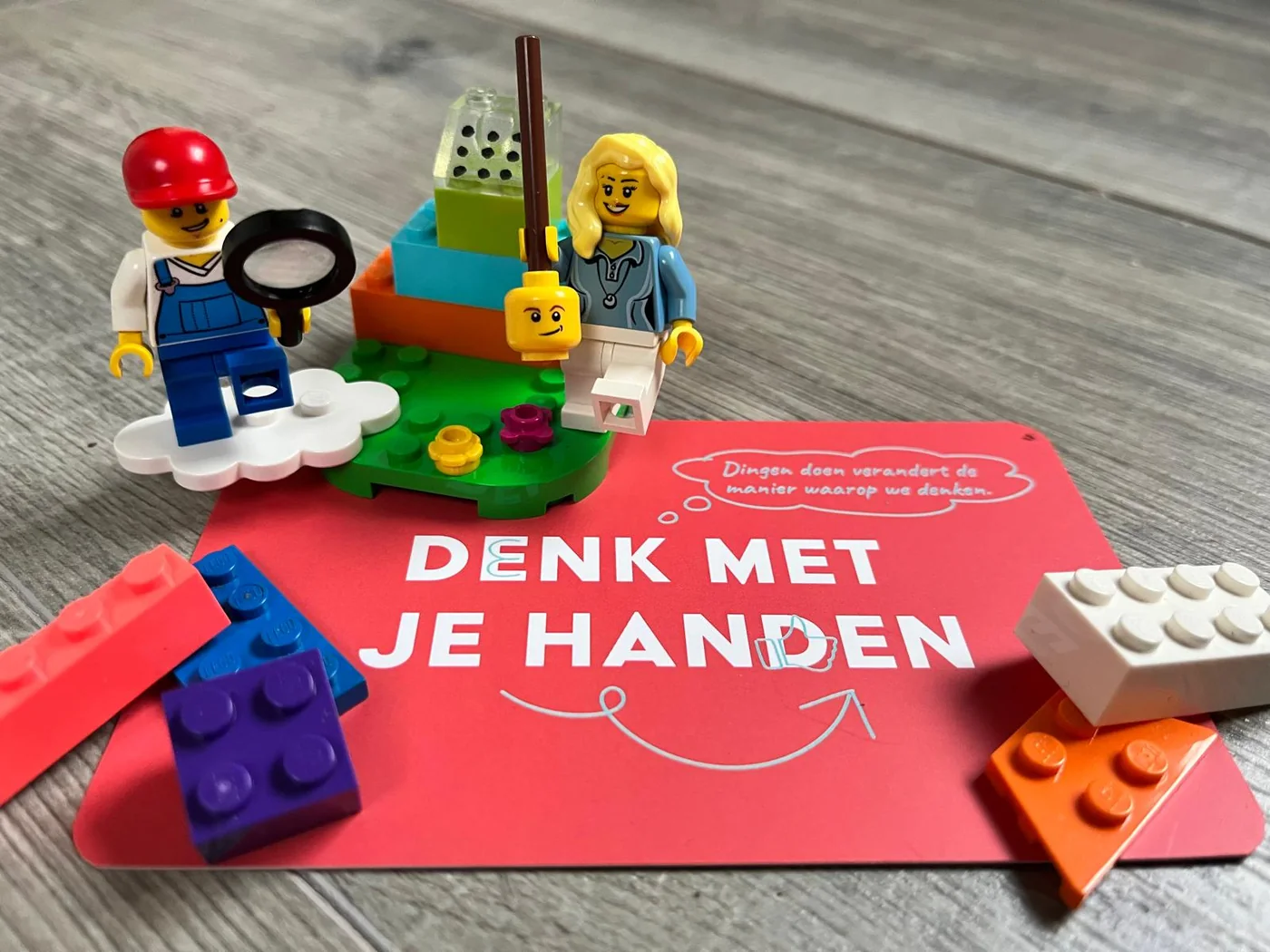 Twee LEGO-poppetjes bij een klein bouwwerk, met losse stenen op een rode kaart met de tekst Denk met je handen