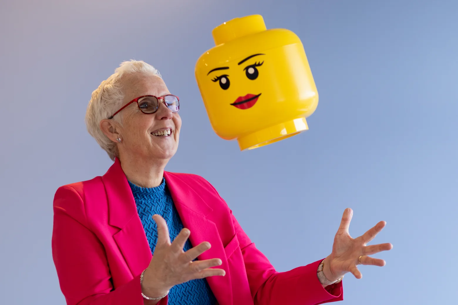 Margot de Jong in een roze pak gooit lachend een groot geel LEGO-hoofd omhoog