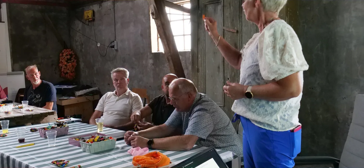 Margot de Jong staat aan het hoofd van een tafel met blokken en legt een bouwopdracht uit aan vier mannen die met hun bouwwerk bezig zijn